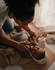 A woman applies lotion to a baby lying on the mushie Star Lovey Blanket. The baby holds a wooden teether, wears a knit hat, and sunlight streams through sheer curtains in the background.