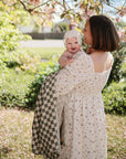 A woman in a floral dress holds a smiling baby wrapped in a mushie Organic Cotton Muslin Swaddle Blanket, standing outdoors beneath blooming branches with sunlight and greenery surrounding them.