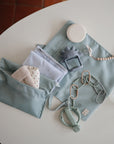 Mushie No-Drop Teether, pacifier clip, and soft cloths in blue pouches on a table.