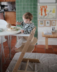 A young child eats from a bowl in a high chair at the dining table, with a Mushie Splat Mat under the chair protecting the floor. The kitchen features green tiles and framed artwork, creating a cozy, homey feel.