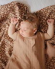 A sleeping baby with curly hair lies on a leopard-print blanket, dressed in a light brown sleeveless outfit. Nearby are the mushie Star Lovey Blanket and a soft star-shaped pillow for added comfort.