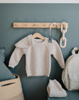 A cozy beige knit baby sweater hangs on a wooden peg rail against a green wall, with a matching beanie, cream backpack, FRIGG Butterfly Anatomical Silicone Pacifier 2-Pack (6-18 Months), toy chain, and cushioned basket with blue pillow nearby.
