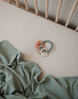 A pastel green blanket lies crumpled on a cream crib sheet, next to a baby teething ring with blue beads and silicone animal shapes. Pale wooden crib bars are visible in the background.
