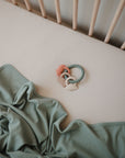 A pastel green blanket lies crumpled on a cream crib sheet, next to a baby teething ring with blue beads and silicone animal shapes. Pale wooden crib bars are visible in the background.