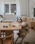 A baby with a knit hat and light outfit plays with the mushie Silicone Baby Rattle Toy while sitting in a high chair at a wooden dining table. The cozy, modern room’s minimalist decor enhances their sensory playtime.