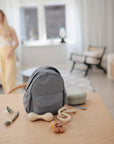 A mushie Silicone Baby Rattle Toy rests on a wooden table in a bright living room alongside a small gray backpack and pacifier clip. In the background, a woman stands near the sofa, slightly out of focus.