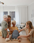 A woman and three children pack a blue backpack in a bright living room. The baby stands on the table, holding a mushie Silicone Baby Rattle Toy made from food-grade silicone, while the others help organize toys.
