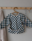 A mushie Long Sleeve Bib in teal and white checks hangs on a wooden peg rack, accompanied by a textured beige cloth, teething toy, pacifier clip, and beaded baby accessory.