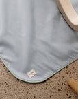 A light blue, water-resistant Mushie Splat Mat with rounded corners and a small MINIMAT tag rests on a terrazzo floor near light wood furniture—ideal as a high chair mat or splat mat.