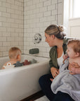 A woman holding a baby in a towel sits by the tub, watching another child play with toy boats. On the counter is mushie Fragrance Free Baby Body Lotion, ideal for keeping delicate skin soft after bath time.