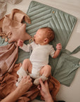 A baby in a white onesie lies on an organic cotton mat, holding the mushie Star Lovey Blanket, surrounded by soft brown and beige fabrics as an adult gently puts socks on their feet.