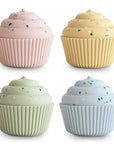 The mushie Mix and Match Cupcake Toy features four pastel cupcakes–pink, yellow, green, and blue–with swirled frosting and sprinkles in ribbed silicone cups, arranged in a 2x2 grid on a white background.