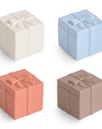 Four small, square Mix and Match Present Toys by mushie—in cream, light blue, coral, and brown silicone with raised bows—are arranged in a 2x2 grid on a white background. Great for creative play and fine motor skill development.