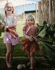 Two girls outdoors holding rhubarb leaves and mushie Linen Pacifier Clip 2-Pack on the grass.