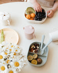 Child’s hands with snacks, plates, and a mushie Dinnerware Fork and Spoon Set on the table.