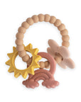 A silicone baby teether with soft beaded loops and attached shapes: a yellow sun, a pink and red rainbow, and a beige star. The brand name mushie is embossed on the loop.