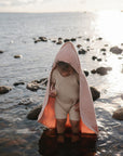 A young child in a beige outfit and pink hooded towel stands among rocky shallows, holding a Mushie FRIGG Rope Natural Rubber Pacifier as sunlight reflects off the water and the shimmering sea glows in the background.