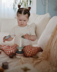A young child with pigtails, dressed in a white shirt and patterned pants, sits on a bed stacking mushie Stacking Rings Toy’s non-toxic rings onto a peg as soft light fills the cozy room.