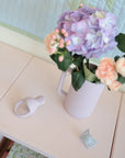 A pastel pink pitcher with purple hydrangeas and pink flowers sits on a light cabinet. Nearby are a Mushie Silicone Fresh Food Feeder and a small blue butterfly-shaped object.