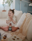 A smiling toddler with pigtails claps her hands beside the mushie Stacking Rings Toy, seated on a white couch. She wears heart-patterned pants, with a soft blanket nearby in the softly lit, cozy room.