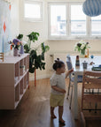 A toddler walks barefoot in a sunlit dining room with a set table, pink chairs, potted plants, and the mushie Stacking Rings Toy placed beside a blue striped lamp hanging from the ceiling.