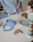 Toddler at table with mushie pastel dishes, bib, cup, and Stacking Rings Toy; adult nearby.