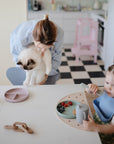A woman holds a fluffy white cat as a toddler with pigtails sits at a table in a bright kitchen, eating fruit and crackers from the mushie Dinnerware Fork and Spoon Set.