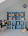 A pastel blue shelf displays colorful mugs beneath a slanted ceiling. Nearby, Mushie Silicone Fresh Food Feeders, a vase with purple and pink flowers, a bowl, a pastel figurine, and a white coffee machine sit on the clean countertop.