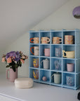 A blue cubby shelf with compartments displays colorful mugs and cups on a white countertop. Nearby are a bouquet of flowers in a pink vase, a mushie Lunchbox, and a coffee machine under a sloped ceiling with a purple wall lamp.