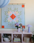 A pastel quilt with geometric patterns hangs above a pink shelf holding books, a vase of flowers, candles, a mushie Stacking Rings Toy, a lavender lamp, and small decor items in a cozy, modern room.