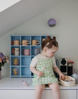 A young girl sits on a white counter under a slanted ceiling, self-feeding with the Mushie Silicone Fresh Food Feeder. Behind her, colorful mugs rest on a blue shelf next to flower-filled vases.