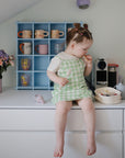 A young girl in a green gingham dress sits on a white counter eating from a beige mushie Lunchbox with built-in compartments. Behind her, mugs sit on a blue shelf and vases with flowers add a decorative touch.