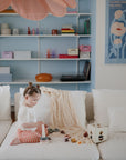 A young child with styled hair sits on a white couch, playing with the mushie Stacking Rings Toy. The pastel-themed living room has shelves with books and decor items, plus a floral poster on the blue wall.