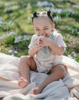 A young child with pigtails sits barefoot outdoors on a light blanket, smiling and holding a snack while wearing her favorite mushie Muslin Bib. Green grass and small purple flowers fill the background.