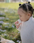 A young child with braided hair enjoys self-feeding outdoors, drinking from the Mushie Silicone Fresh Food Feeder, surrounded by blurred purple flowers.