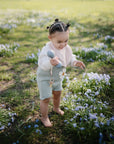 A toddler with styled hair stands barefoot on grass among purple flowers, wearing a light sweater and shorts, holding the mushie Silicone Baby Rattle Toy and looking down curiously in a bright outdoor setting.
