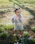 A toddler stands barefoot on grass with small flowers, holding and chewing a mushie Silicone Baby Rattle Toy. She wears a white sweater and green overalls with a daisy design, enjoying a pleasant sensory experience.
