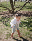 A toddler with braided hair walks barefoot on grass, holding a mushie Silicone Baby Rattle Toy in each hand. She wears a white sweater, green shorts, and a pink bib. A tree with bare branches stands in the background.