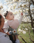 A woman holds a baby in a flower-themed outfit and pink bib as the child reaches for white tree blossoms, enjoying spring outdoors while clutching the mushie Silicone Baby Rattle Toy.