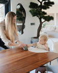 A woman with long blonde hair hands a cup to a toddler in a high chair at a wooden dining table, where Mushie First Feeding Baby Spoons are nearby in a modern, plant-filled living space with neutral decor.