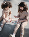 Two young girls in matching light brown outfits sit on concrete, playing with the Mushie Wooden Puzzle. They appear focused and engaged as they use this educational toy to build fine motor skills together.