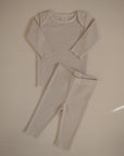 The mushie Two Piece Set features a light beige ribbed long sleeve shirt and pants made from breathable, soft fabric, perfect for children. Shown laid flat on a neutral background.