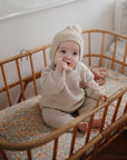 A baby in a beige knit outfit and hat sits in a wooden crib with a floral mattress, holding the mushie Lamb Figurine Teether and looking to the side.