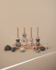 The Mushie Wooden Multi Shape Stacker features pastel rings and rods on a neutral beige background. Some rings are stacked, others scattered, encouraging fine motor skills, all set in soft natural lighting for a calm educational atmosphere.
