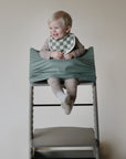 A smiling blonde toddler sits in a high chair, wearing a striped outfit and green-and-white checkered bib. The mushie Multi-Use Cover is wrapped around the chair for gentle security. Background is plain and light-colored.