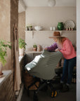 A woman in a pink sweater and hat leans over a stroller covered with a green mushie Multi-Use Cover in a sunlit room decorated with potted plants, shelves, and rustic accents.