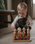 A young child in overalls sits on a wooden floor, stacking colorful shapes on the Mushie Wooden Multi Shape Stacker, an educational toy that helps develop fine motor skills. A patterned cushion is nearby and soft light fills the room.