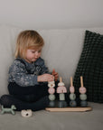 A blonde toddler sits on a light grey sofa stacking the Mushie Wooden Multi Shape Stacker to build fine motor skills, with small animal figurines nearby and a dark green cushion beside her.