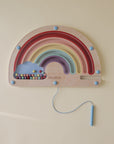The mushie Wooden Magnetic Rainbow Maze features colorful arches, a blue cloud, and pastel beads. Kids use a handled blue string to guide beads on the pale yellow surface, encouraging fine motor skills and screen-free play.
