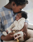 A young child in a blue and white checkered sweater kisses a sleeping baby wearing mushie’s Zipper Pajama made from cozy organic cotton, as they sit by a window soaked in gentle natural light.